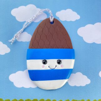 Handmade polymer clay chocolate egg in a cup. Shaped in a classic egg shape, the top half includes a faux chocolate egg, with a criss cross pattern. The bottom half features the egg cup - blue and white striped with a cute smiling face - black glass bead eyes, a big smile and rosy cheeks. Measuring 7cm by 4.5cm.