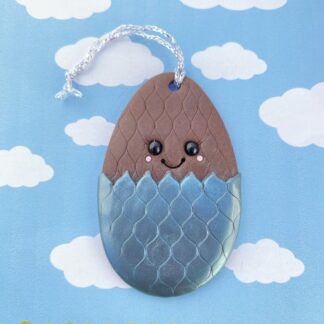 Handmade polymer clay chocolate Easter egg tree decoration. Made using the classic egg shape, the chocolate egg has a criss-cross pattern with the bottom half complete with a foil effect. Finished with a cute smiling face, black glass bead eyes, big smiling face and rosy cheeks. Measuring 7cm by 4.5cm.