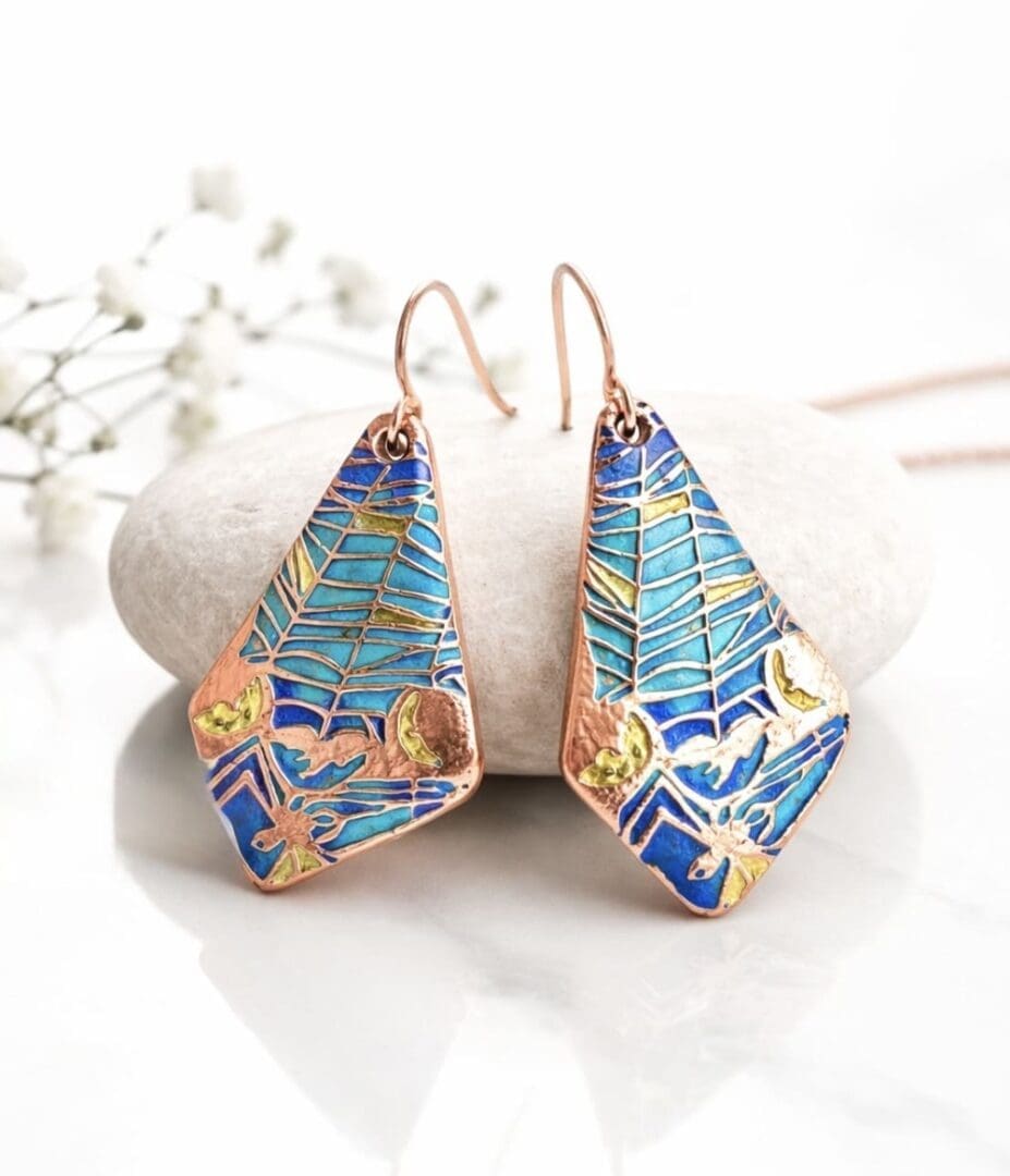 A pair of copper drop earrings in a domed extended triangle shape, textured a nd hand painted in turquoise shades of metallic paint. They have rose gold plated ear wires and displayed on a white background with a white gift box above it.