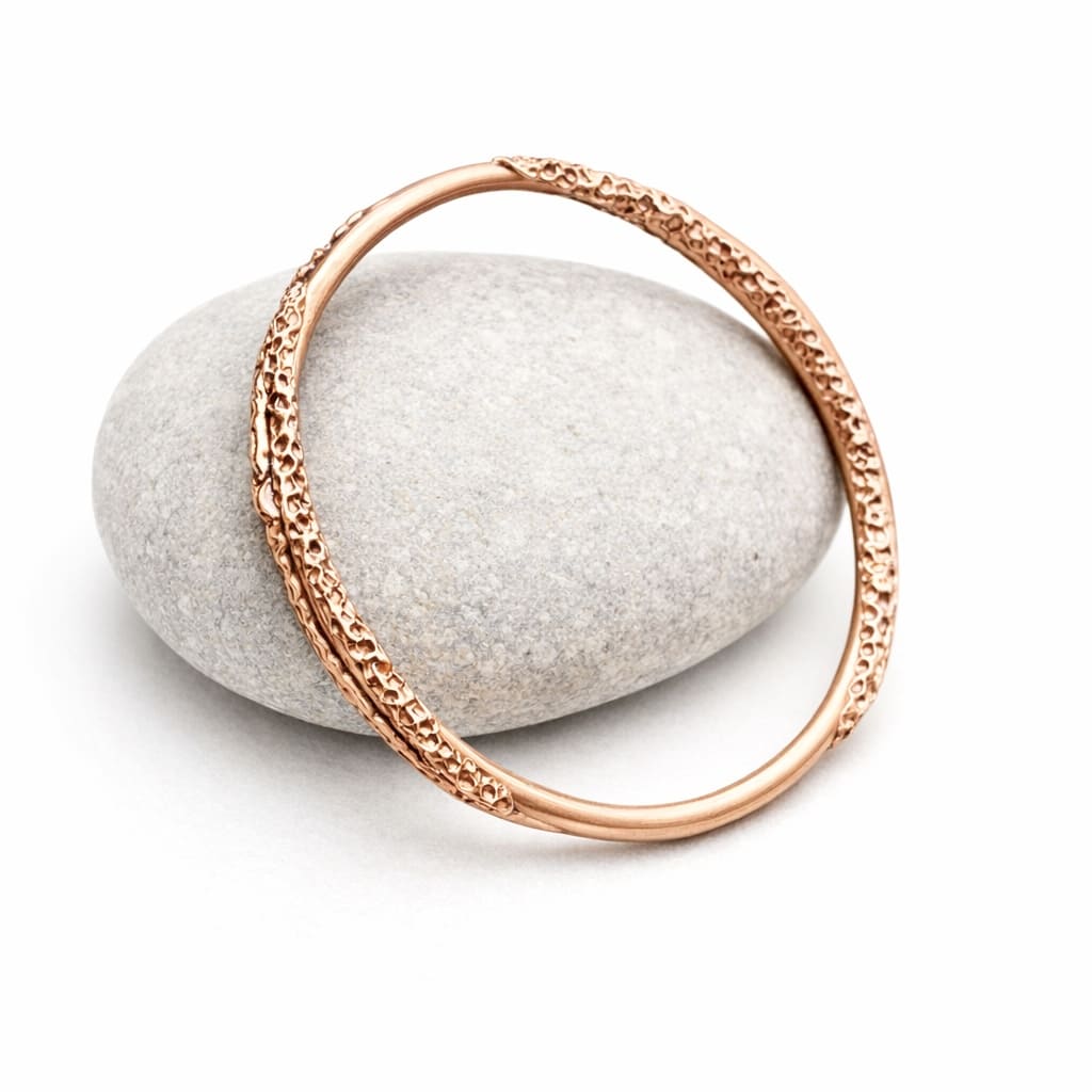 A handcrafted copper bangle with a unique textured pattern aroung the outer edge. The bangle is displayed on a white background resting on a light coloured stone.