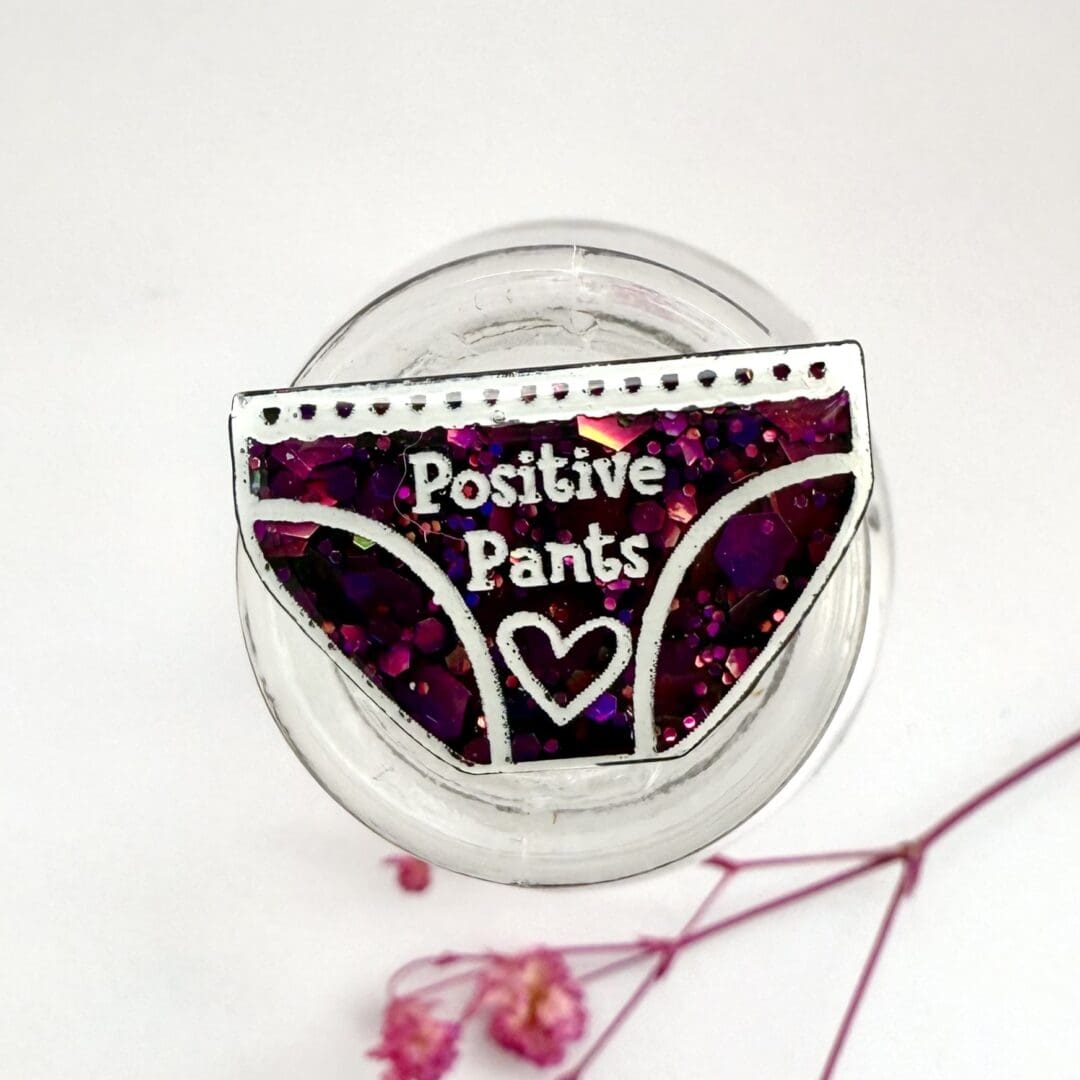 Dark pink glitter positive pants pin badge with brass coated pin is displayed on a glass jar with a white background and pink flowers.