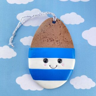 Handmade polymer clay boiled egg in a cup. Shaped in a classic egg shape, the top half includes a light brown speckled egg, coloured with pastels. The bottom half features the egg cup - blue and white striped with a cute smiling face - black glass bead eyes, a big smile and rosy cheeks.