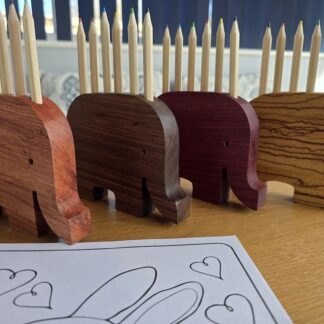 Handcrafted wooden elephant pencil holders in exotic woods including zebrano, walnut, purpleheart and African rosewood, displayed with colouring pencils on a desk
