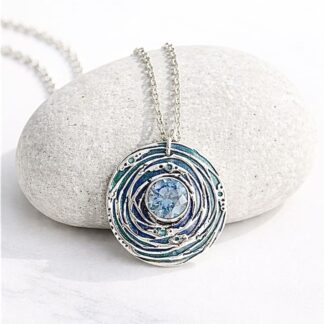 A handcrafted fine silver round shaped pendant that has a 8mm. blue cubic zirconia stone set in the centre. The pendant has been hand- painted in between the design pattern, in blue metallic points, the necklace has a sterling silver chain and is displayed resting on a stone against a white background.
