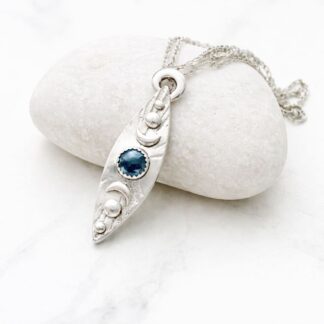 A fine silver long and narrow pendant with a 10mm. hematite stone. It is displayed on a white background resting on a light coloured stone.