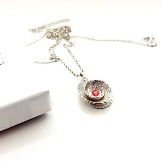 A small fine silver coiled textured pendant with a 5mm red cubic zirconia stone in the centre, with a sterling silver chain. Displayed on a white background.