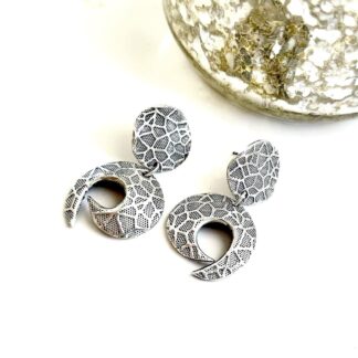 A pair of fine silver oxidised round stud and drop earrings with a textured pattern and organic half moon shape with a small circle and a larger one hanging from it. They have stainless steel ear posts and are displayed on a washed white wood background.