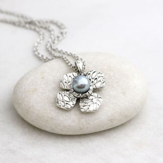 A fine silver flower shaped pendant with a patterened surface and a small light grey pearl in the centre. Displayed on a pale grey stone.