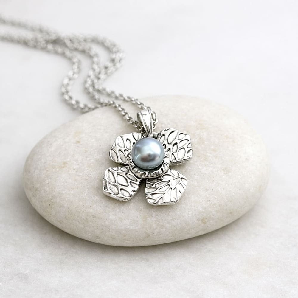 A fine silver flower shaped pendant with a patterened surface and a small light grey pearl in the centre. Displayed on a pale grey stone.