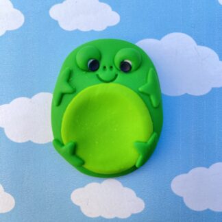 A green clay frog worry stone with big eyes and a smile sits in front of a blue sky with white clouds.