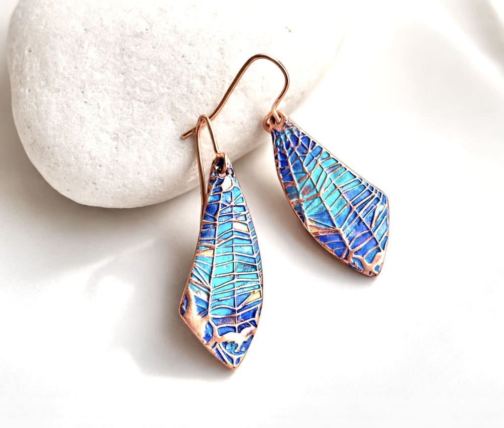 A pair of handcrafted copper drop earrings with a spider web design hand-paonted in metallic blues and turquoise paints. They are in a wing shape and are displayed resting on a white stone with a white background.