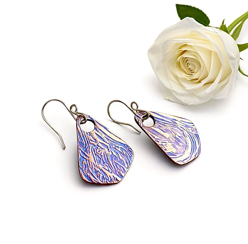 A pair of handcrafted triangle shaped copper drop earrings with a large hole for the sterling silver ear wires. Hand-painted blue metallic paint, these textured earrings are displayed on a white background.