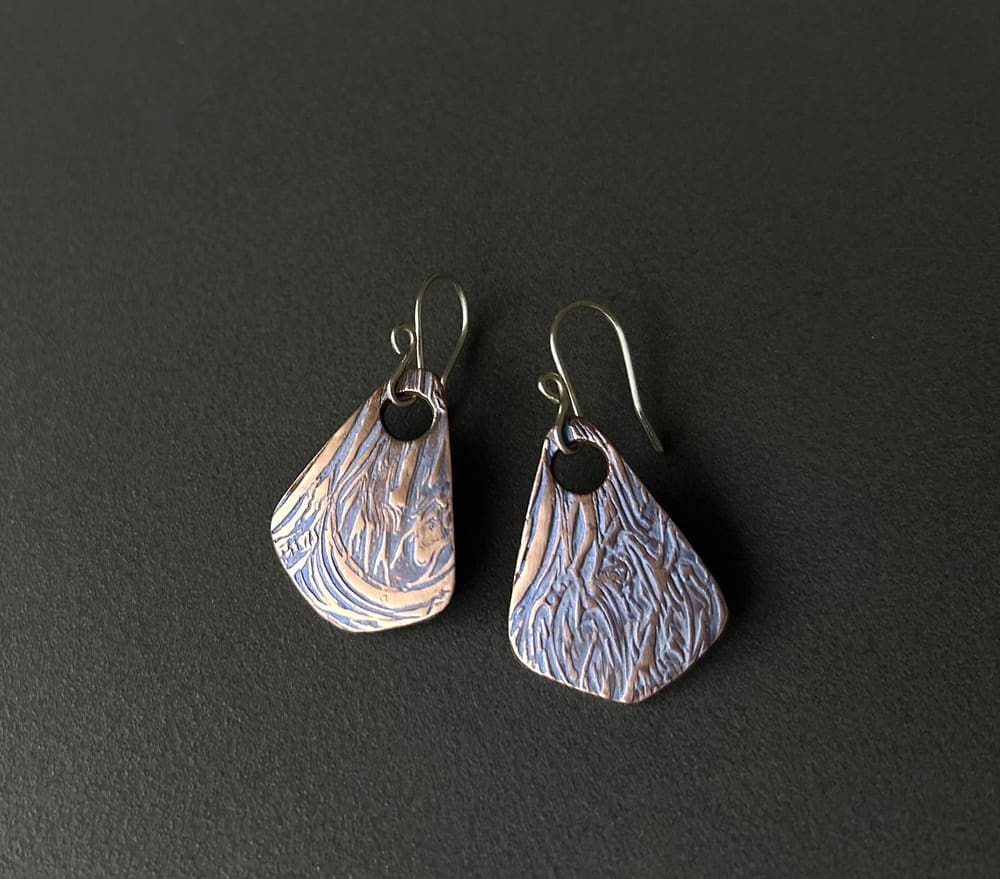 A pair of handcrafted triangle shaped copper drop earrings with a large hole for the sterling silver ear wires. Hand-painted blue metallic paint, these textured earrings are displayed on a dark grey background.