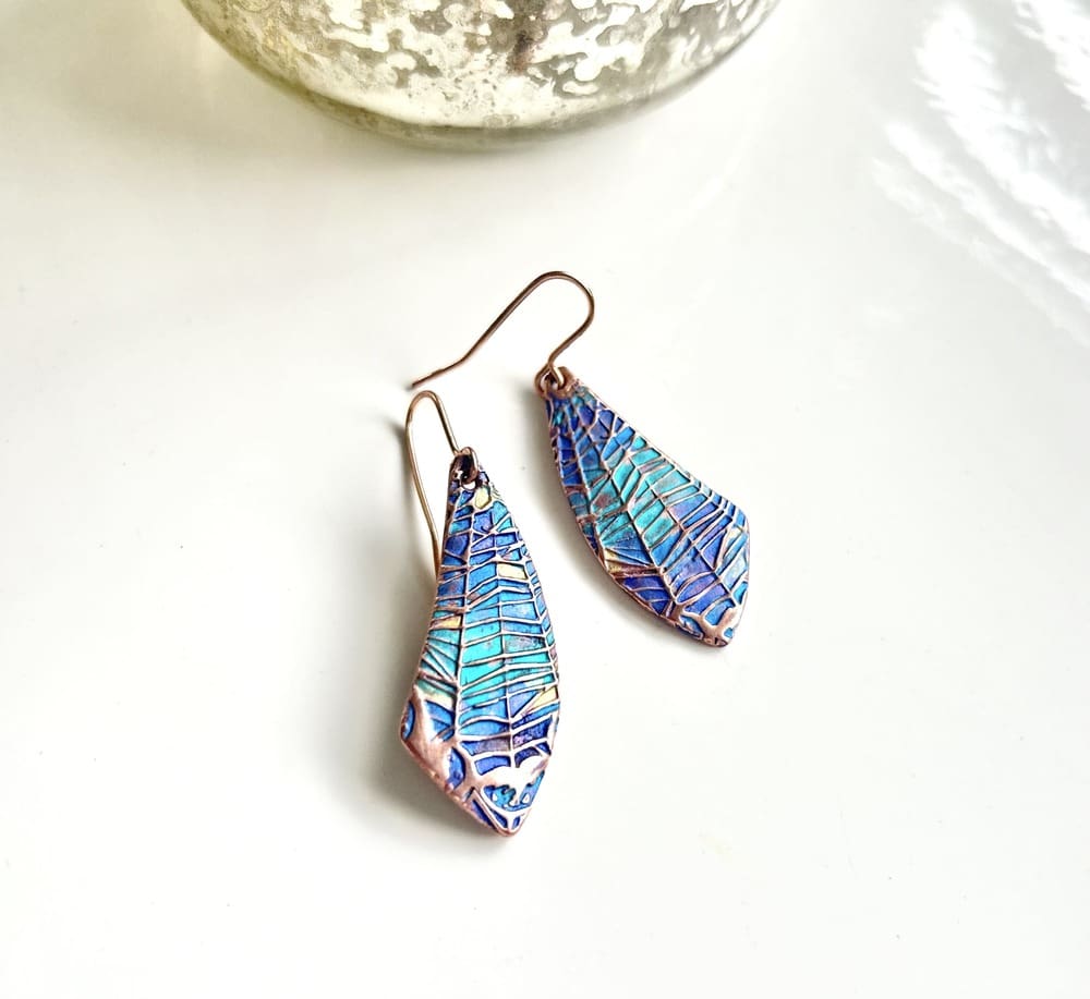 A pair of handcrafted copper drop earrings with a spider web design hand-paonted in metallic blues and turquoise paints. They are in a wing shape and are displayed on a white background.