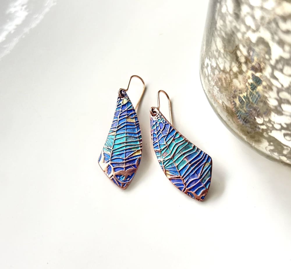 A pair of handcrafted copper drop earrings with a spider web design hand-paonted in metallic blues and turquoise paints. They are in a wing shape and are displayed on a white background.
