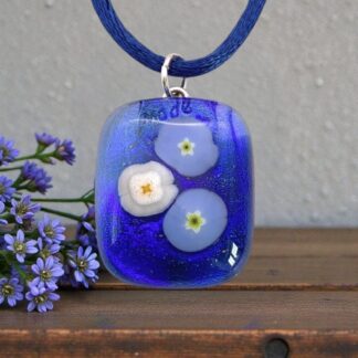 Glass pendant with blue and white flower designs on a blue cord, placed on a wooden surface beside small purple flowers.