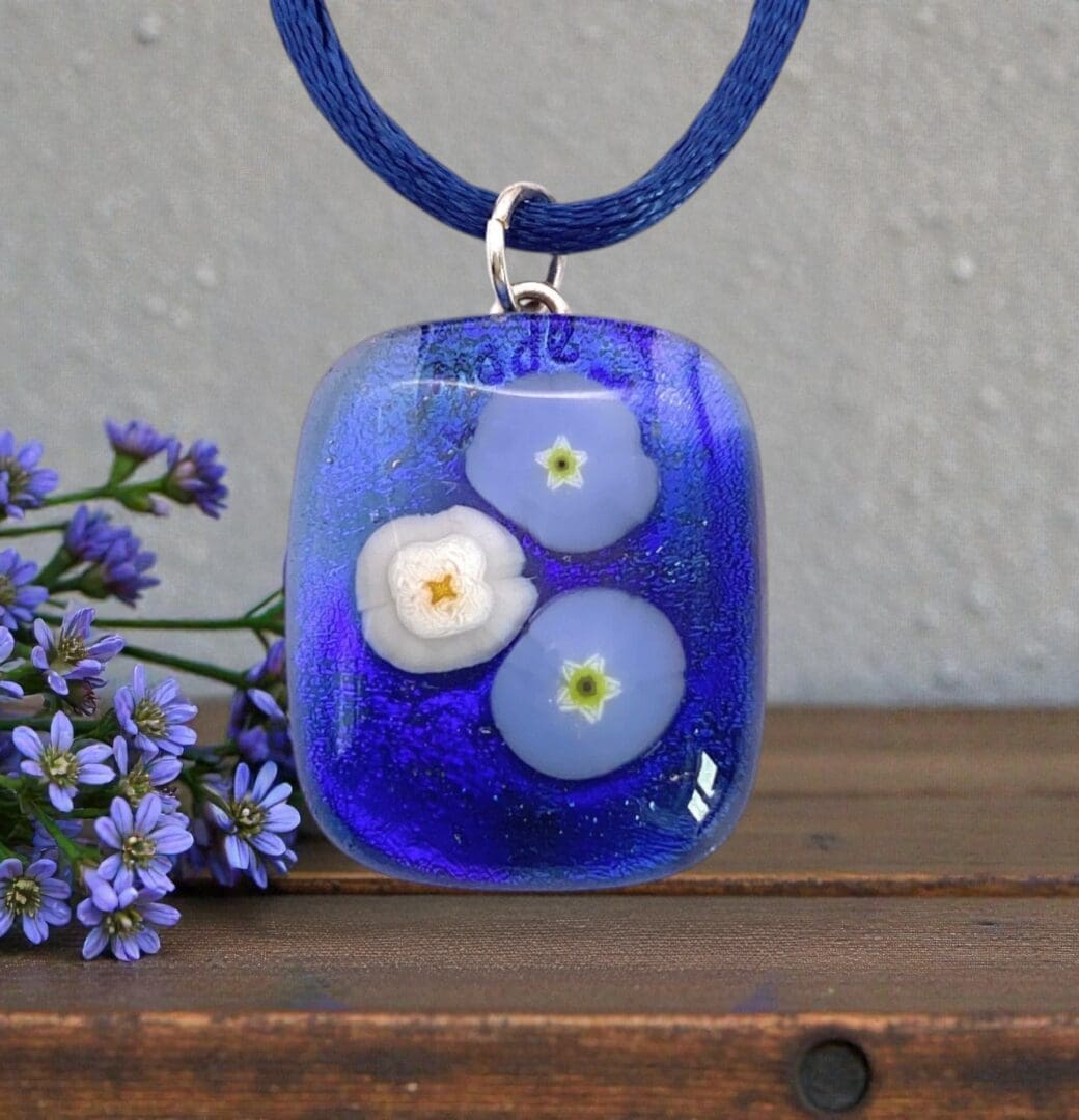 Glass pendant with blue and white flower designs on a blue cord, placed on a wooden surface beside small purple flowers.