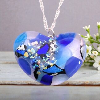 Heart-shaped glass pendant with blue, purple, and silver shards, hanging on a silver twisted chain, next to small white flowers.