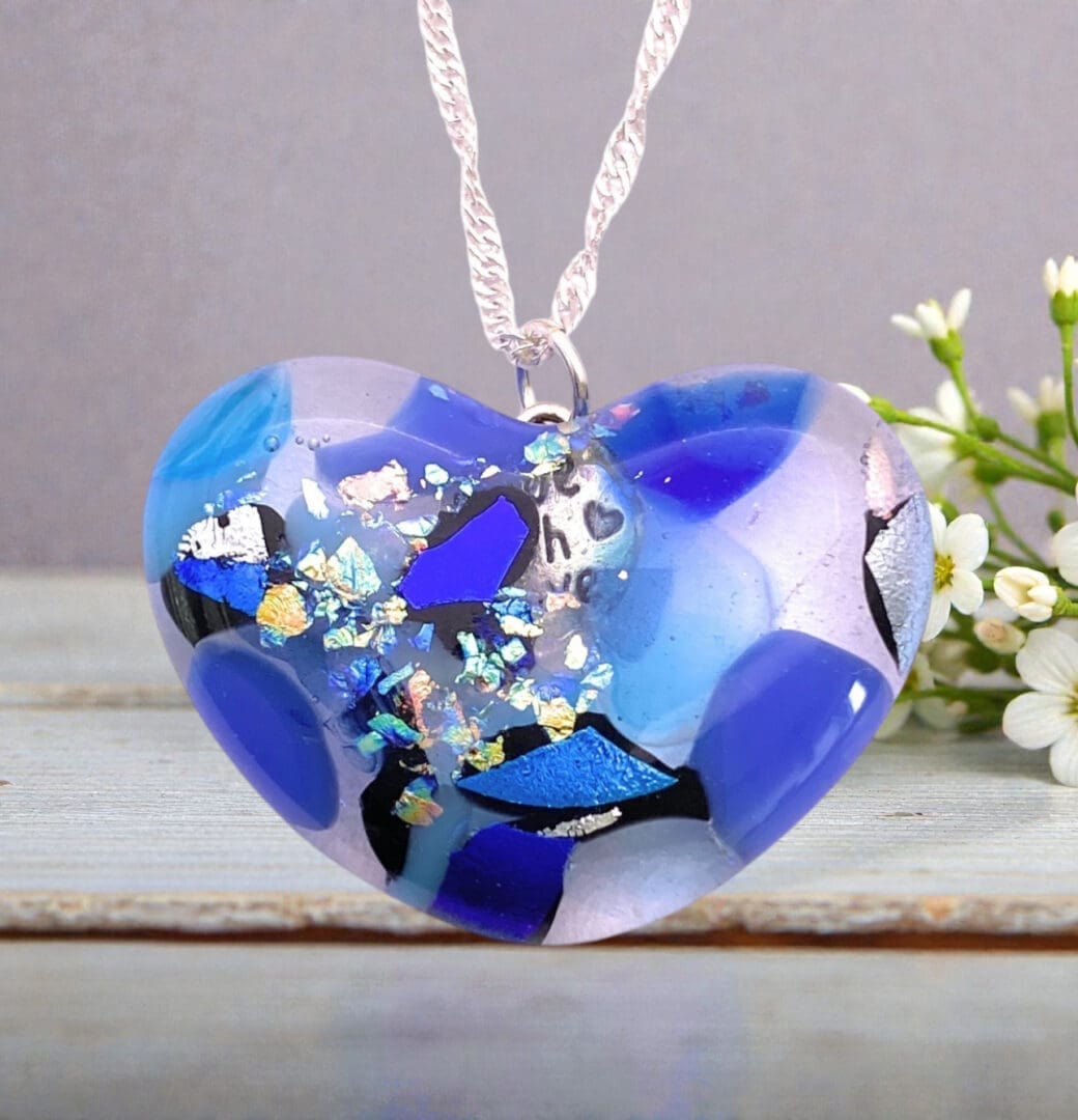 Heart-shaped glass pendant with blue, purple, and silver shards, hanging on a silver twisted chain, next to small white flowers.