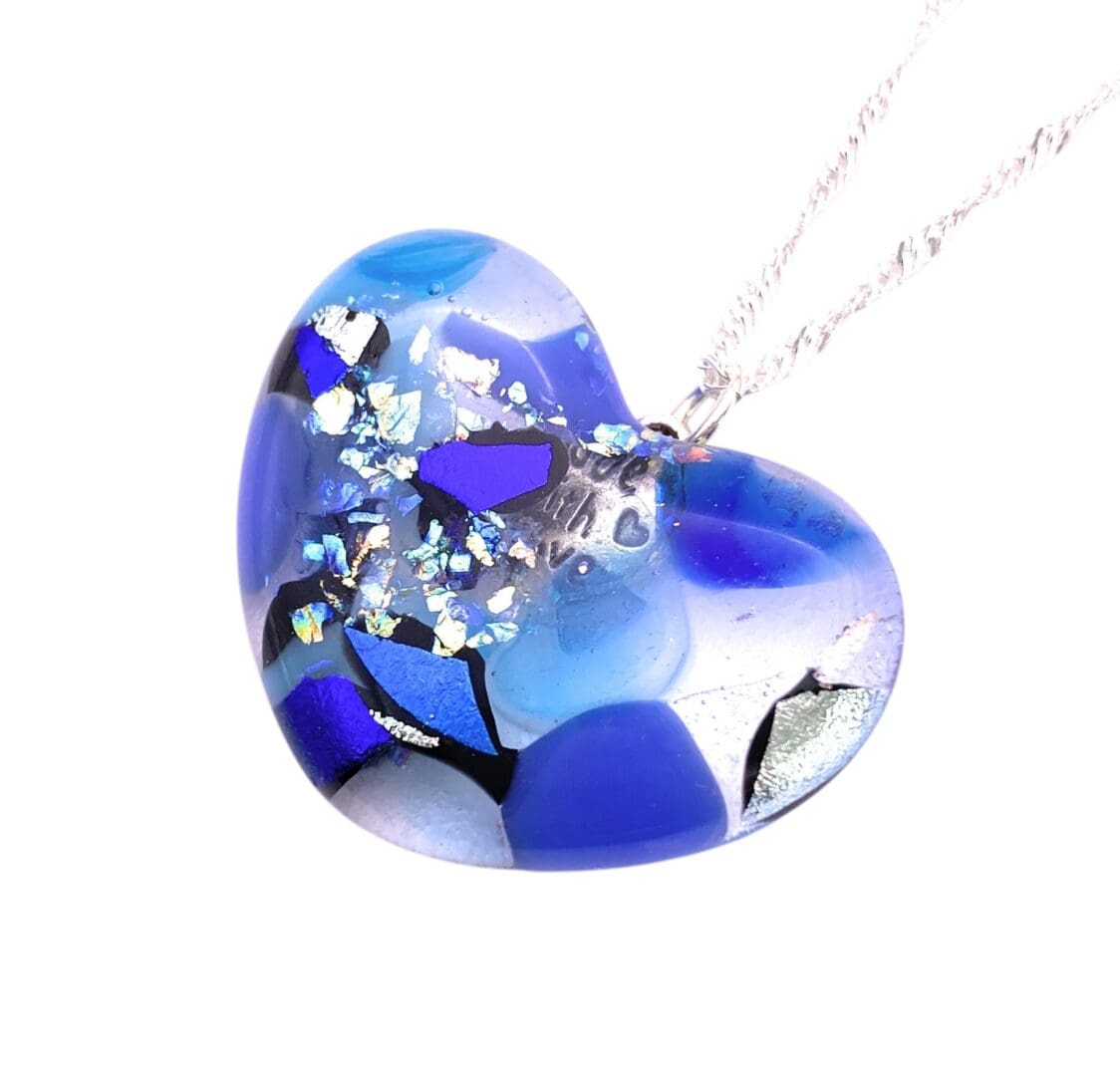 A blue heart-shaped glass pendant with silver flakes and varying blue and black patterns. It hangs from a delicate silver chain, exuding elegance.