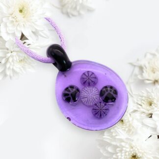 A purple glass pendant featuring intricate designs, with a black bail and a lavender cord, surrounded by white flowers.
