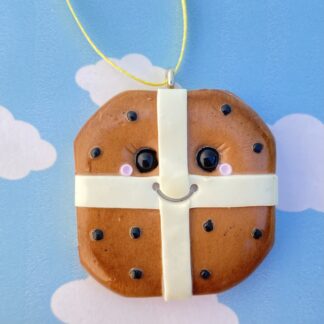 Handmade polymer clay hot cross bun tree decoration, square in shape with rounded corners, hanging from a yellow ribbon. The bun includes a light coloured base with roughened sides to represent the dough, with a brown topping, light coloured cross and little black raisins. Finished with a beautiful smiling face and rosy cheeks, all with a glossy shine.