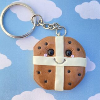Handmade polymer clay hot cross bun keyring, square in shape with rounded corners, hanging from a key chain. The bun includes a light coloured base with roughened sides to represent the dough, with a brown topping, light coloured cross and little black raisins. Finished with a beautiful smiling face and rosy cheeks, all with a glossy shine.