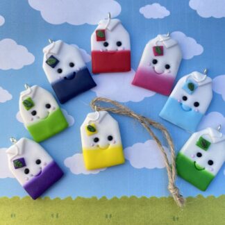 A collection of eight handmade kawaii tea bag pot tags with smiling faces and colored bottoms in pink, purple, blue, navy, yellow, green, and red, set against a backdrop of a sky with clouds and a grassy field.