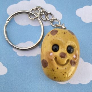 Handmade polymer clay potato keyring. Made using a blend of yellow, browns and shimmering gold. With additional brown spots and pastel "dirt". Features a cute smiling face, black glass bead eyes a little brown spot nose, big smile and rosy cheeks. measuring 3cm by 2.5cm, Overall length 8.5cm