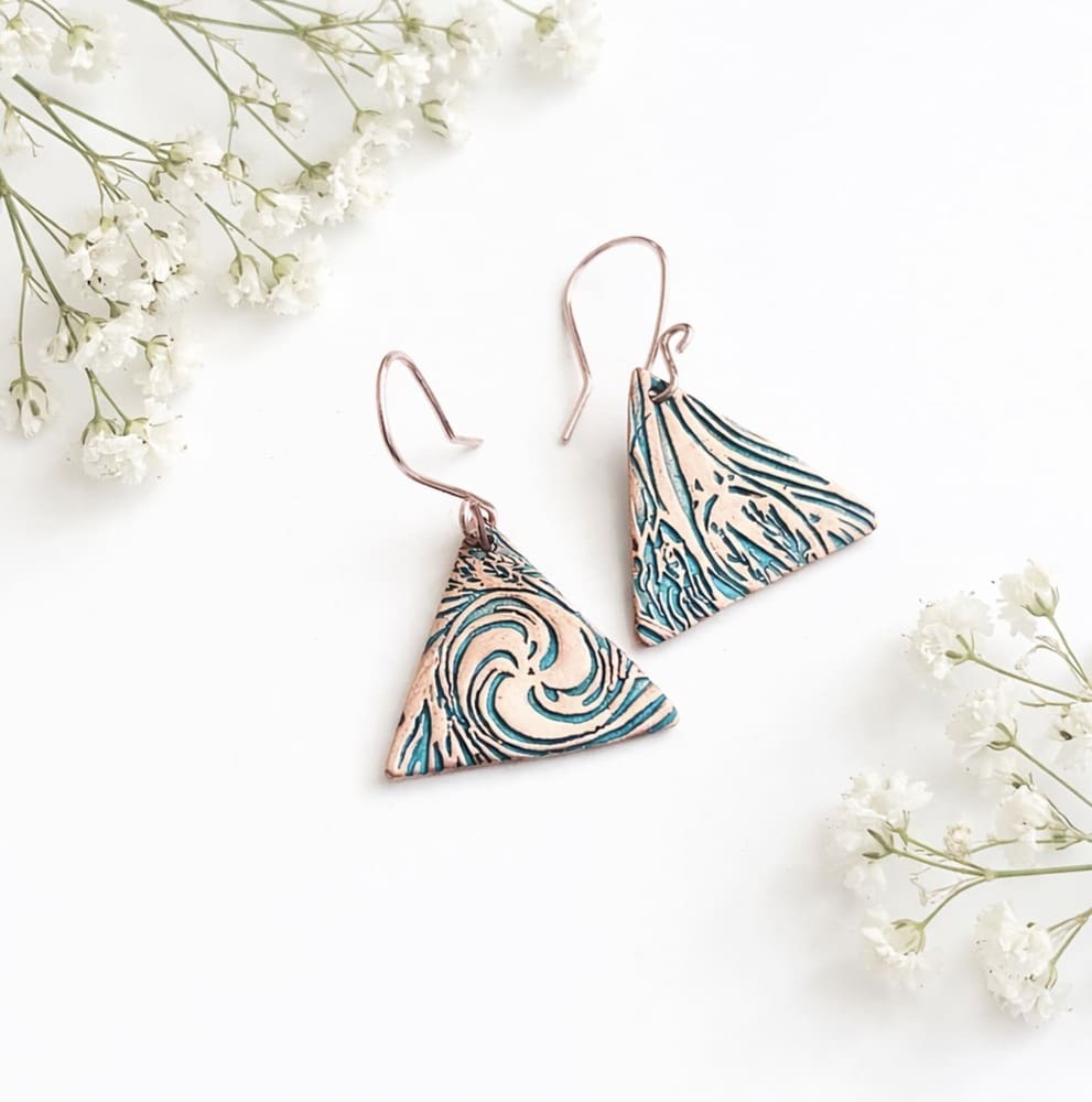A pair of copper triangle shaped drop earrings that have a textured pattern and have sterling silver ear wires. They are displayed on a white background with a few babys breath flower sprigs.