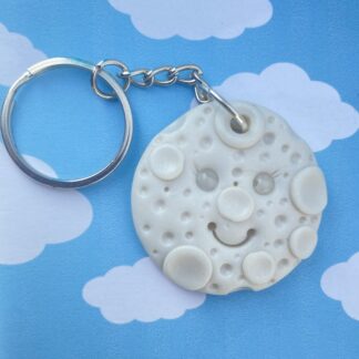A handmade keychain featuring a smiling moon character with a textured surface and attached to a silver keyring. The background is a whimsical scene of a blue sky with white clouds.