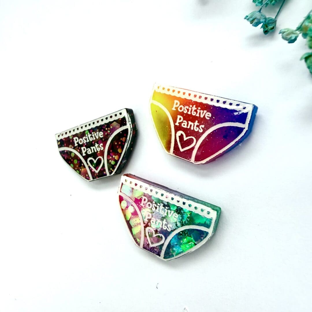 Side view of positive pants badges, neon rainbow, mermaid mix (pink and teal), dark pink glitter) displayed on a white background with blue flowers.