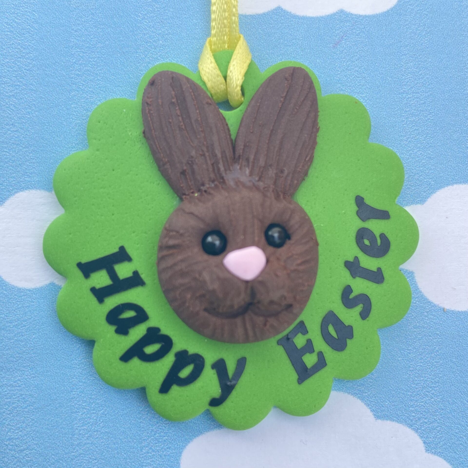 Handmade polymer clay bunny rabbit tree decoration. Each bunny features a sculpted brown face with long upright ears, a pink nose, sparkling black bead eyes, and a cheerful smile, all set on a vibrant green base and finished with a sunny yellow ribbon for easy hanging. Includes the writing - happy easter 2026, to show the option of personalisation