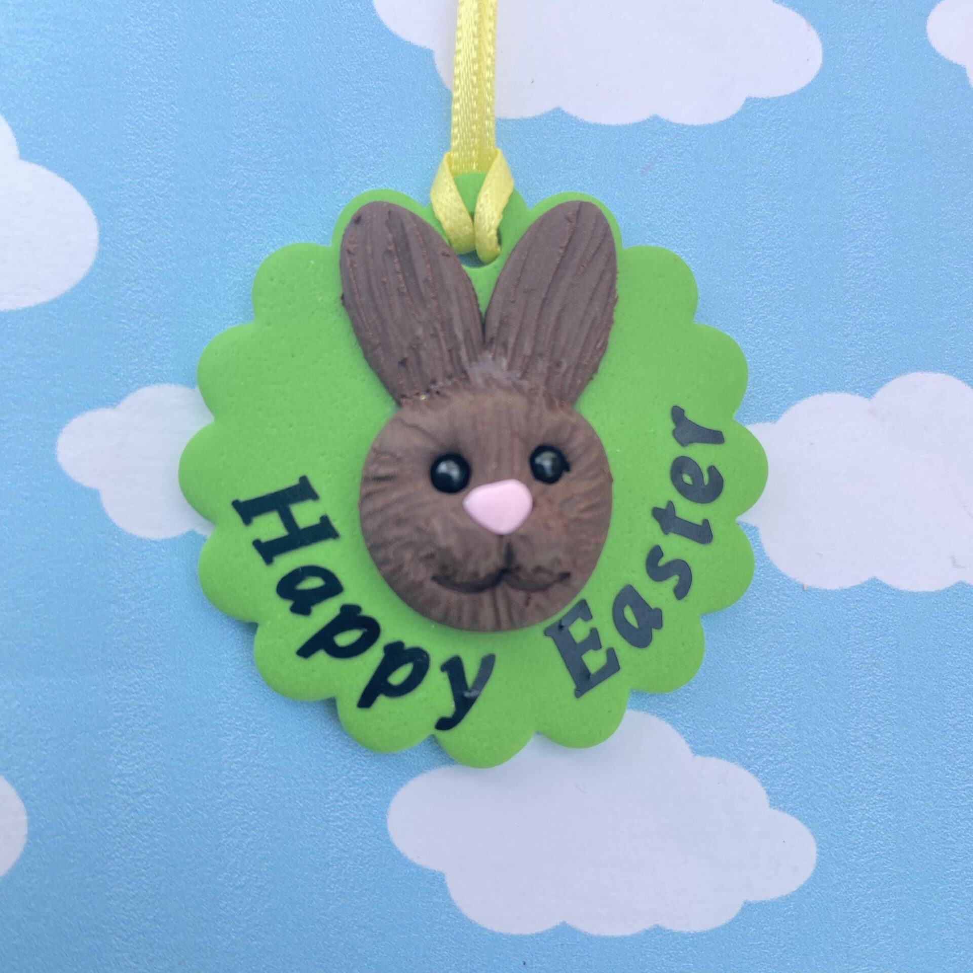 Handmade polymer clay bunny rabbit tree decoration. Each bunny features a sculpted brown face with long upright ears, a pink nose, sparkling black bead eyes, and a cheerful smile, all set on a vibrant green base and finished with a sunny yellow ribbon for easy hanging. Includes the writing - happy easter 2026, to show the option of personalisation