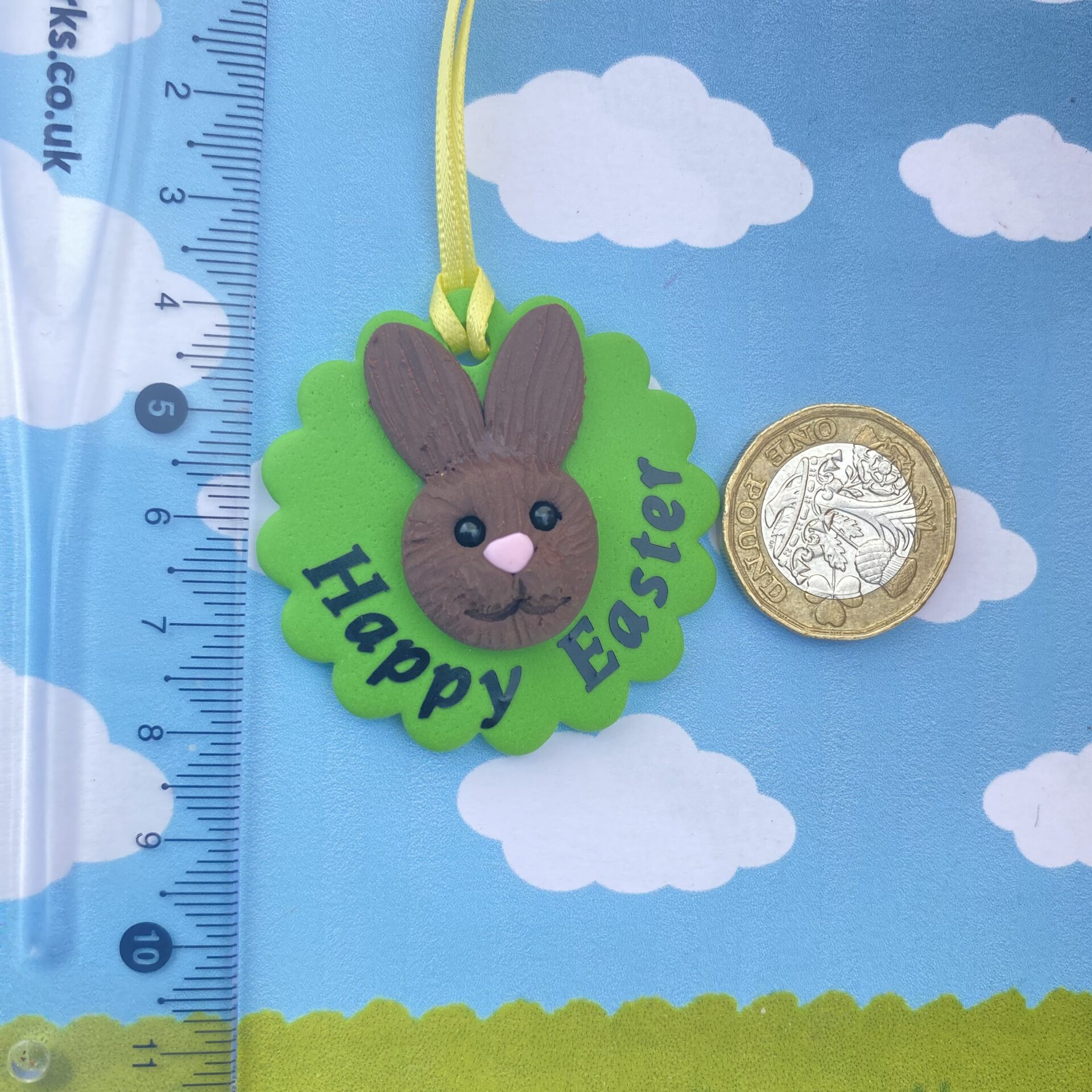 Handmade polymer clay bunny rabbit tree decoration. Each bunny features a sculpted brown face with long upright ears, a pink nose, sparkling black bead eyes, and a cheerful smile, all set on a vibrant green base and finished with a sunny yellow ribbon for easy hanging. Includes the writing - happy easter 2026, to show the option of personalisation