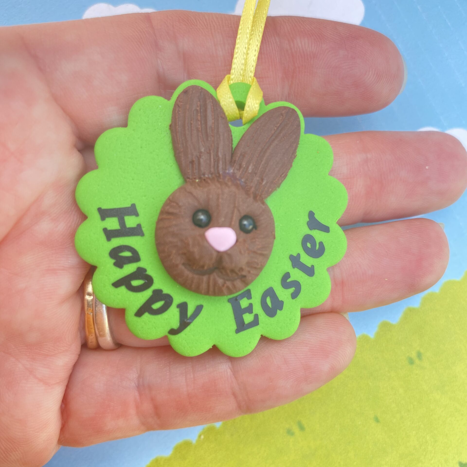 Handmade polymer clay bunny rabbit tree decoration. Each bunny features a sculpted brown face with long upright ears, a pink nose, sparkling black bead eyes, and a cheerful smile, all set on a vibrant green base and finished with a sunny yellow ribbon for easy hanging. Includes the writing - happy easter 2026, to show the option of personalisation
