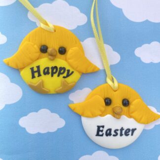 Two handmade polymer clay chick, easter decorations. Both with cute faces, small glass bead eyes and a gold beak. The left one has a bright yellow belly and little gold feet, with "happy" in black writing and the one on the right is sat in a white shell and peaking out, with Easter wrote on it.