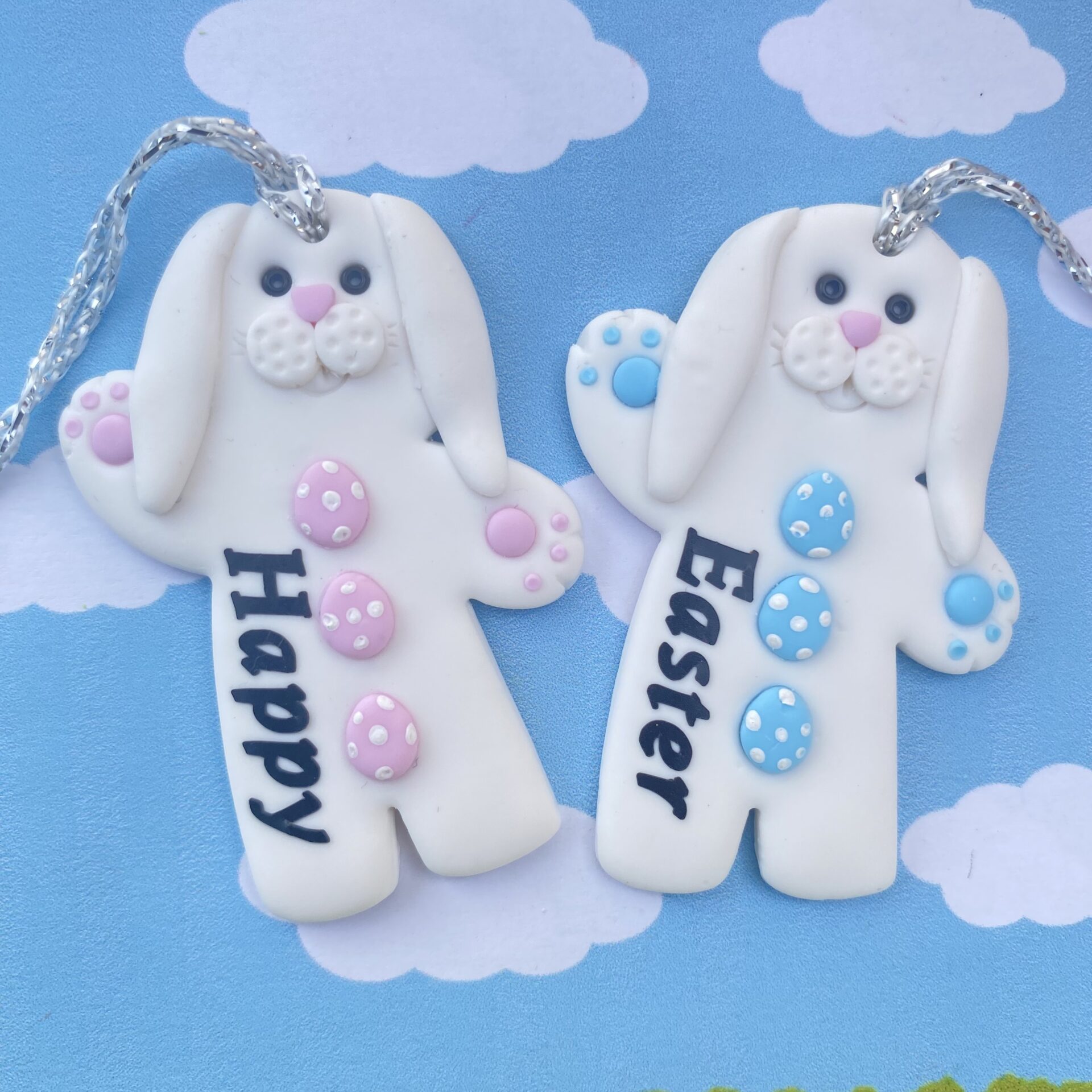 Two white bunny-shaped ornaments with pastel-colored eggs on their chests, set against a blue cloudy sky background. The left ornament in pale pink reads "Happy" in black script, and the right in pale blue reads "Easter" in black text.
