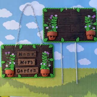Two handmade polymer clay wooden effect signs, showing the choice of a hanging chain or garden stake, with a green vine border effect and small leaves. The sign says "Nan's Herb Garden" with a small terracotta pot on each side with a smiling face and green leaf plants and coloured flowers sprouting from them.