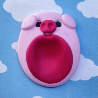 A pink clay pig worry stone with black eyes and a dark pink snout and belly sits against a blue sky with white clouds.