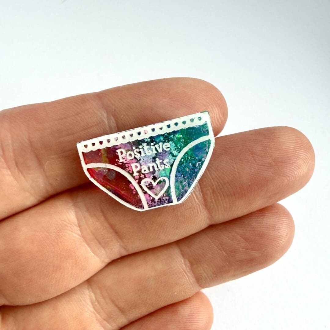 Pink and teal positive pants pin badge with holographic glitter displayed on a hand for scale