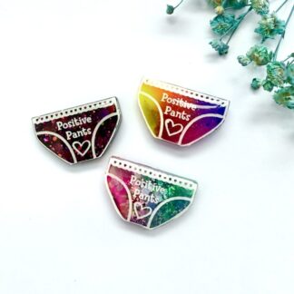 Selection of positive pants badges, neon rainbow, mermaid mix (pink and teal), dark pink glitter) displayed on a white background with blue flowers.