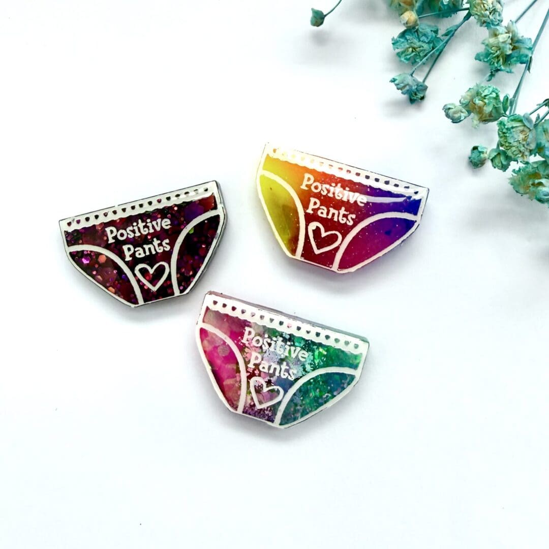 Selection of positive pants badges, neon rainbow, mermaid mix (pink and teal), dark pink glitter) displayed on a white background with blue flowers.