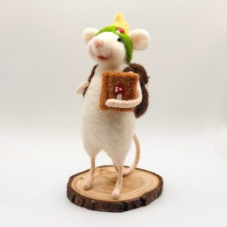 Needle felt white mouse wearing school backback in brown, holding tan felt book featuring a mushroom logo. Wearing green and yellow hat with mushroom logo. Bag contains wooden pencils and paper scrolls.