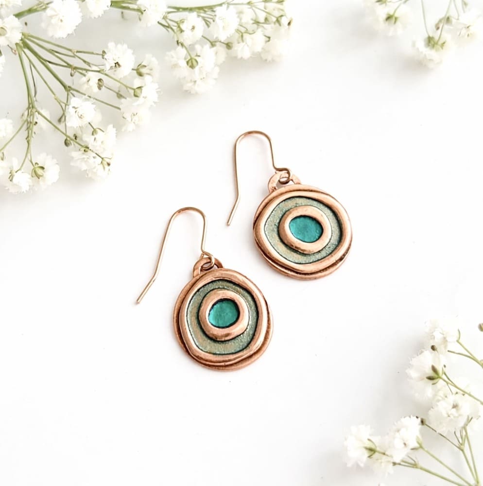A pair of small round copper drop earrings with copper ear wires, the double circle design has been hand-painted, the outer circle is pale green metallic paint and the centre circle is kingfisher metallic paint. The earrings are displayed on a white background.