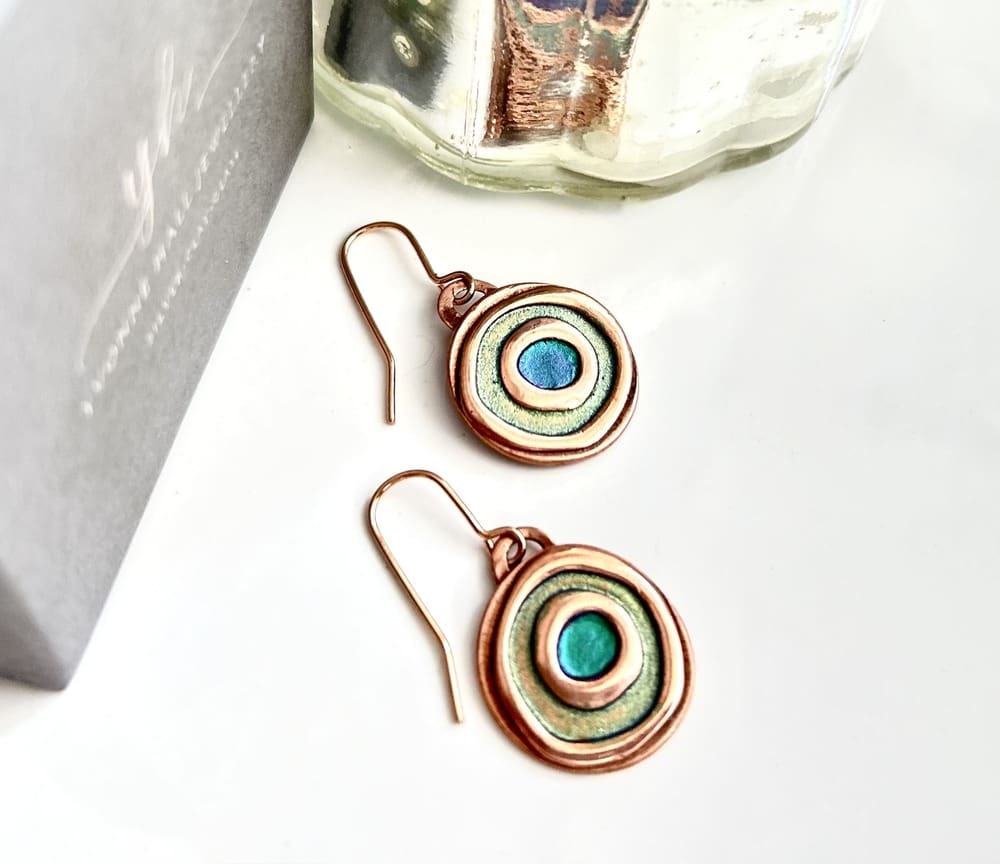 A pair of small round copper drop earrings with copper ear wires, the double circle design has been hand-painted, the outer circle is pale green metallic paint and the centre circle is kingfisher metallic paint. The earrings are displayed on a white background.