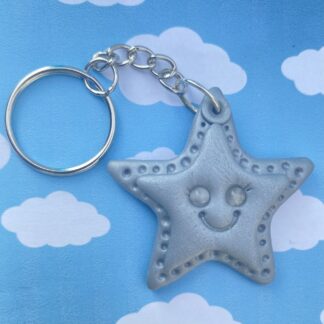 A shimmering silver star-shaped keychain charm with a smiley face, attached to a silver keyring, set against a blue sky with white clouds.