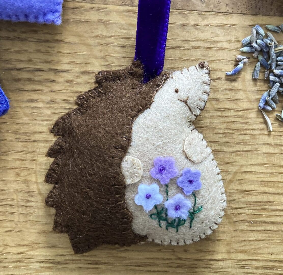 Close up of my lavender filled handmade felt hedgehog, the hedgehog is holding purple flowers and there is a purple velvet ribbon so it can be hung up in a wardobe.