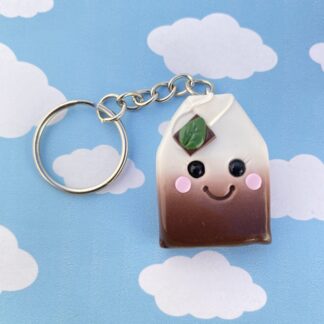 Handmade polymer clay tea bag keyring. Made using a blend of white and brown for the tea effect. With the string tag tucked on top with a green leaf and a cute smiling face, including black glass bead eyes and rosy cheeks.