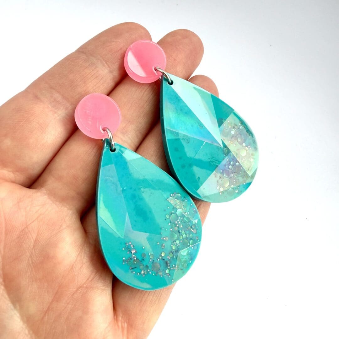 Teal holographic teardrop earrings hung on pink Glow-in-the-dark acrylic stud toppers with surgical steel posts and backs, shown on a hand for size.
